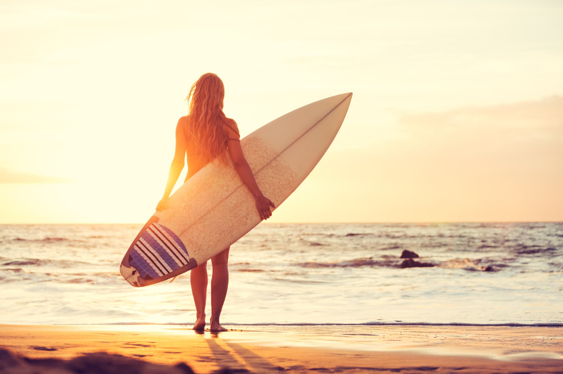 A surfer standing at the shoreline holding a surfboard at sunset